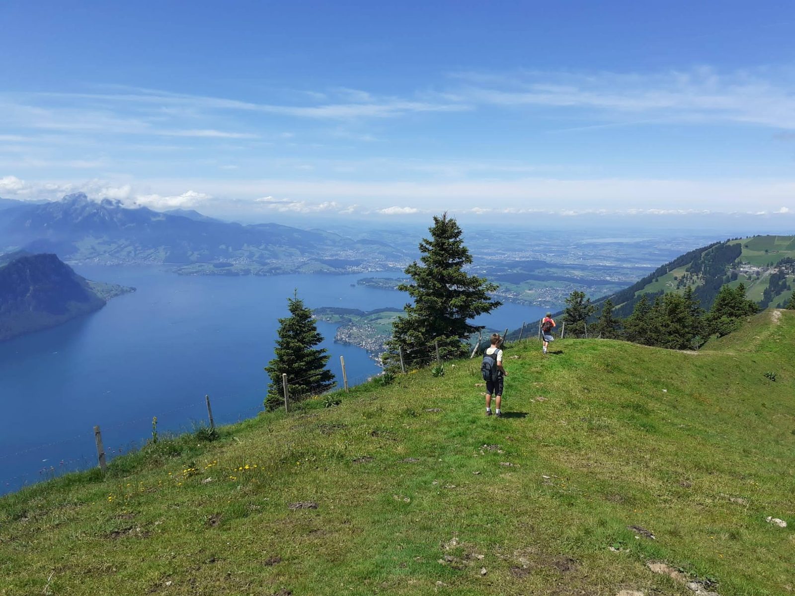 Two young hikers walking down a grassy ridge above Lake
              Lucerne, mountains and haze stretching to the horizon.