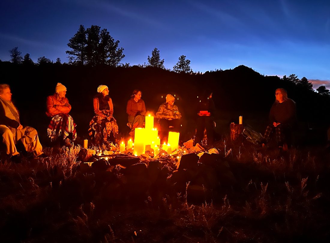 The group sitting in a circle at dusk around a ring of candles, mountains behind
