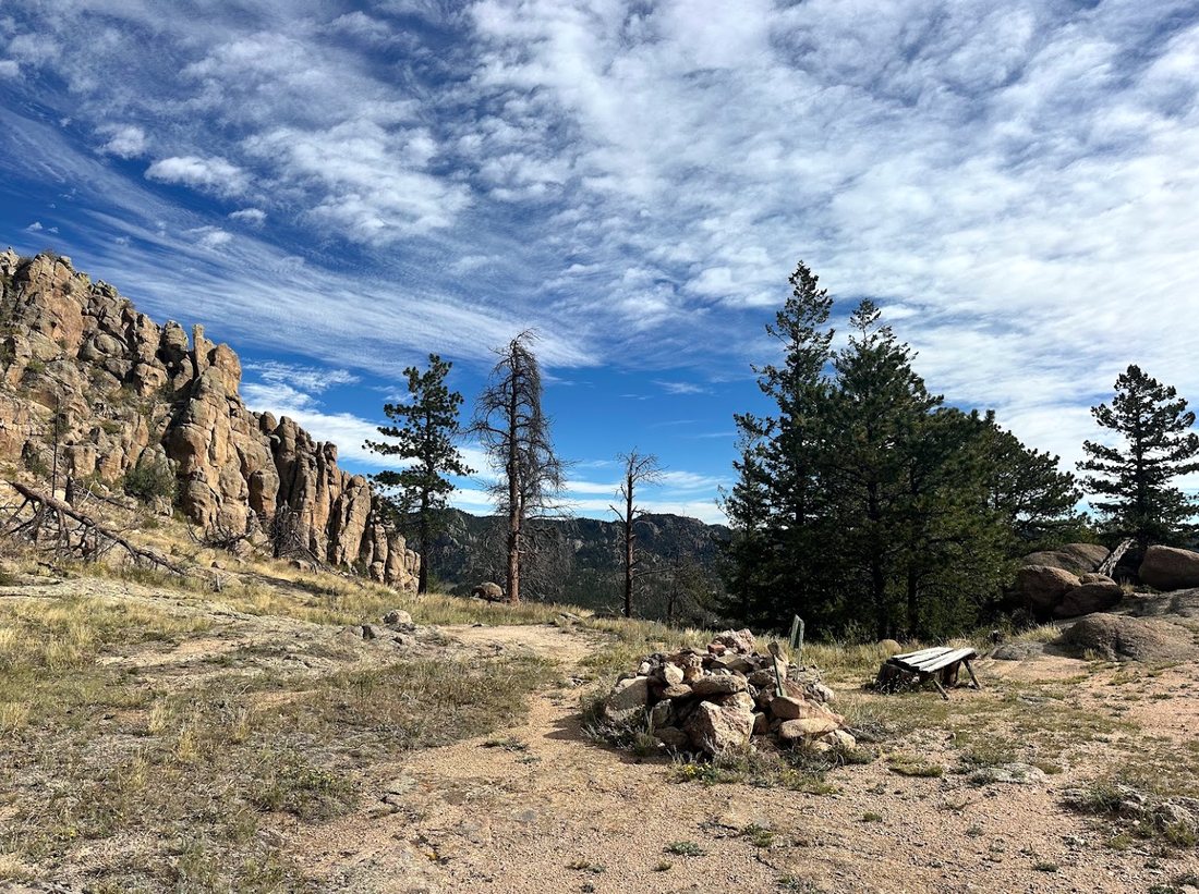 A rocky ridge and pines under a wide Colorado sky, with a cairn beside the trail